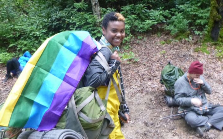 a backpacking student with a pride flag draped over their backpack smiles at the camera